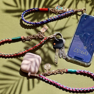 Puede incluir: Un conjunto de tres correas de teléfono trenzadas de colores con herrajes dorados. Las correas están unidas a una funda de teléfono transparente, una funda blanca para AirPod y un dije dorado. La funda del teléfono tiene un reloj digital que muestra las 19:60.