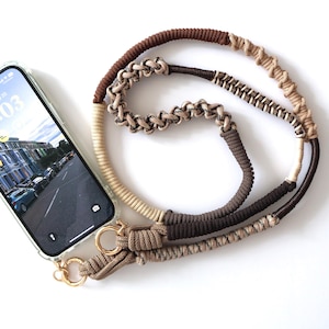 Könnte beinhalten: Ein Telefoncase mit einem Bildschirm, der eine Straßenszene zeigt, verbunden mit einem mehrfarbigen Seil-Handyband. Das Band hat Braun- und Beigetöne mit goldfarbener Hardware. Der Bildschirm zeigt die Uhrzeit 10:03 an.