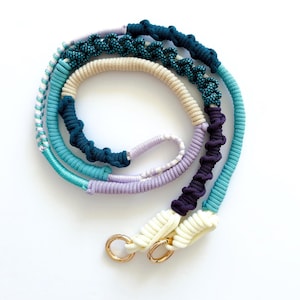 May include: A colorful dog leash featuring a variety of rope textures and colors. The leash includes teal, light blue, cream, purple, and dark blue sections. Gold-colored metal rings are attached to the cream-colored end.