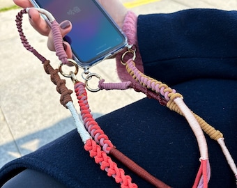 May include: A clear phone case with a colorful macrame phone strap. The strap features a braided design in shades of pink, coral, brown, and white, attached with gold-colored rings. The phone screen displays Chinese characters.