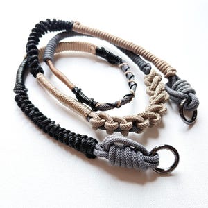 Puede incluir: Tres collares de cuerda hechos a mano en negro, gris y beige. Cada collar presenta un diseño anudado y un cierre de anillo metálico. Las cuerdas están dispuestas sobre una superficie blanca, mostrando las diferentes texturas y colores.