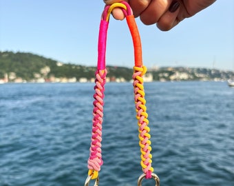 May include: A vibrant phone strap in a braided style, showcasing pink, yellow, and orange colours. Gold-toned rings attach the strap to a clear phone case. The background displays water and a blue sky, creating a bright contrast.
