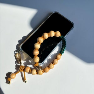May include: A clear phone case with a wooden bead and green string phone strap. The strap is attached to the phone case with a gold metal clasp.