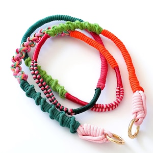 Puede incluir: Tres collares de cuerda coloridos en verde, rosa y naranja, con anillos de metal dorado. Los collares están hechos de cuerda gruesa y trenzada y están dispuestos sobre un fondo blanco. Los colores son vibrantes y llamativos.