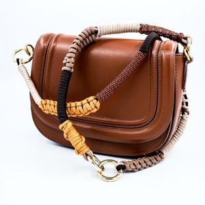 May include: A brown leather handbag with a unique, multi-colored braided strap. The strap features beige, black, tan, and brown sections. Gold-colored metal rings connect the strap to the bag. The bag has a curved flap and a glossy finish.
