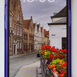 May include: A purple phone case with a gold ring on the bottom. The phone screen shows a photo of a street in a European city with brick buildings and flowers. The time on the phone screen is 18:08.