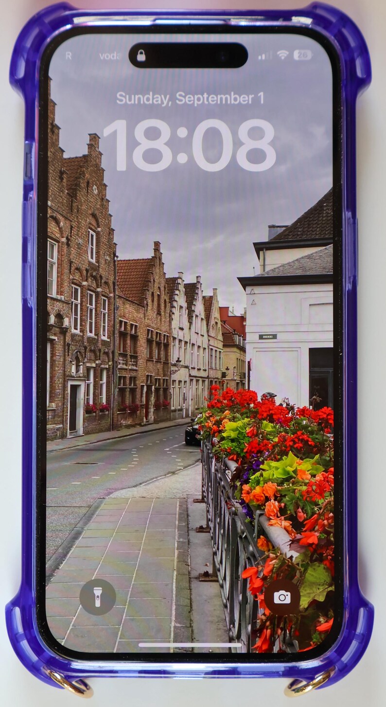 May include: A purple phone case with a gold ring on the bottom. The phone screen shows a photo of a street in a European city with brick buildings and flowers. The time on the phone screen is 18:08.