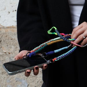 May include: A smartphone case with a colorful braided lanyard. The lanyard features a variety of colors, including blue, green, red, and yellow. The phone case is silver and has metal rings for attaching the lanyard.