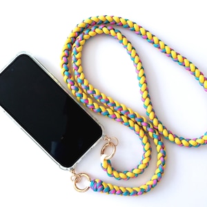 Puede incluir: Un cordón trenzado para teléfono amarillo, azul y rosa con herrajes dorados. El cordón está sujeto a un smartphone en una funda transparente. La pantalla del teléfono es negra. El cordón está hecho de un material grueso y duradero.