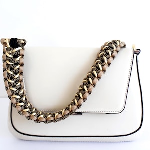 Puede incluir: Un bolso de hombro color crema con un cuerpo rectangular estructurado y ribetes negros en los bordes. El bolso incluye una correa gruesa y decorativa tejida con una combinación de cordones negros, beige y dorados metálicos.