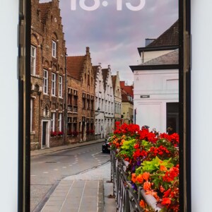 May include: A screenshot of an iPhone displaying a photo of a street in a European city. The time is 18:15 and the date is Sunday, September 1. The photo shows a narrow street lined with brick buildings and colorful flowers.
