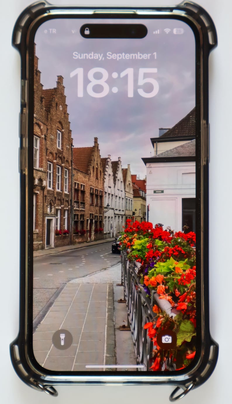 May include: A screenshot of an iPhone displaying a photo of a street in a European city. The time is 18:15 and the date is Sunday, September 1. The photo shows a narrow street lined with brick buildings and colorful flowers.