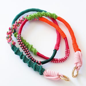 May include: A collection of colorful rope necklaces in shades of green, pink, orange, and red. Each necklace features a unique design, including braided and knotted sections, with gold-colored metal rings.