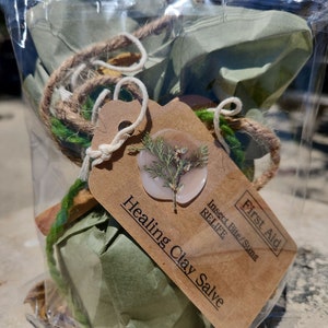 May include: A small, green, paper-wrapped package with a brown tag that reads "Healing Clay Salve" and "First Aid Insect Bite/Stings Relief". The tag has a pink wax seal with a dried green plant inside.