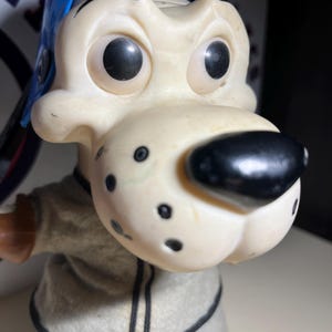 May include: A white plastic toy dog wearing a blue baseball cap and a grey and black jersey. The dog has large black eyes and a black nose.