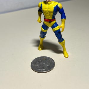 May include: A yellow and blue plastic action figure of a superhero with brown hair. The figure is standing on a white surface next to a US quarter.