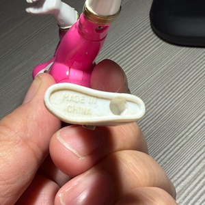 May include: A close-up of a pink and white action figure with gold accents. The figure's foot is held up, revealing the text "MADE IN CHINA" on the sole. The figure is partially visible, with a focus on the leg and foot.