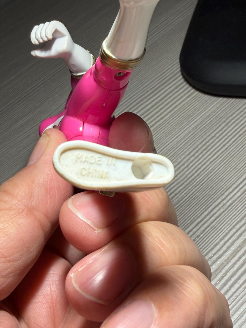 May include: A close-up of a pink and white action figure with gold accents. The figure's foot is held up, revealing the text "MADE IN CHINA" on the sole. The figure is partially visible, with a focus on the leg and foot.