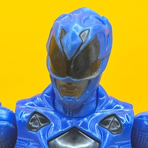 May include: A close-up of a blue action figure's head and upper body. The figure has a blue helmet with black eye coverings and a blue chest piece with black accents. The background is a solid yellow.