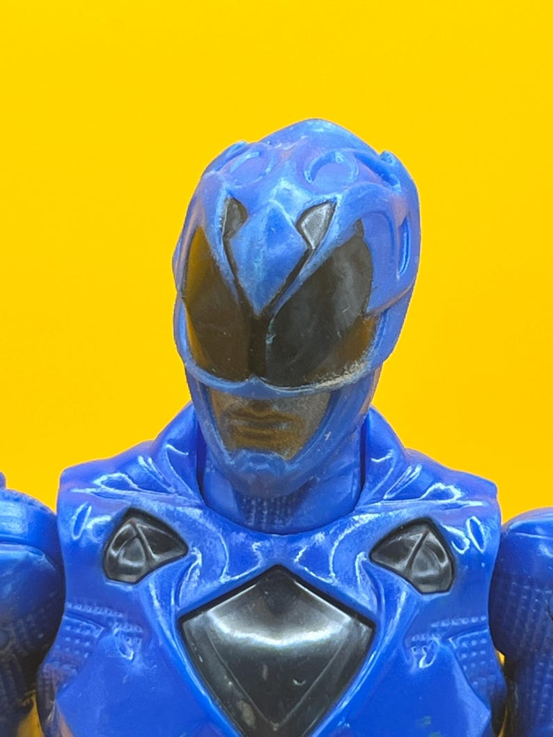 May include: A close-up of a blue action figure's head and upper body. The figure has a blue helmet with black eye coverings and a blue chest piece with black accents. The background is a solid yellow.