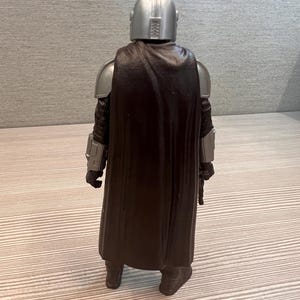 May include: A silver helmeted action figure of a character from a science fiction series, viewed from the back. The figure has silver shoulder armor, black gloves, and a dark brown cape. The figure is standing on a light-colored surface.