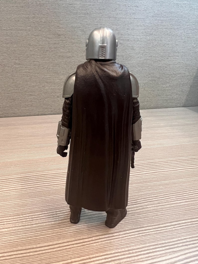 May include: A silver helmeted action figure of a character from a science fiction series, viewed from the back. The figure has silver shoulder armor, black gloves, and a dark brown cape. The figure is standing on a light-colored surface.