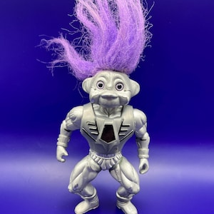 May include: A silver-colored troll doll with purple hair. The doll has a muscular physique and is wearing a silver outfit with a red accent. The troll is standing on a blue background.