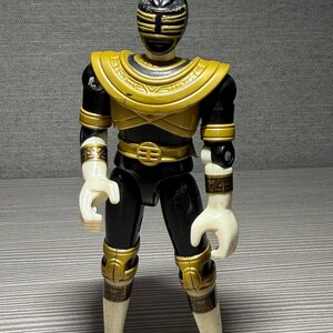 May include: A black and gold Power Ranger action figure. The figure features a black helmet with gold detailing, a gold chest plate, and gold accents on the arms, legs, and belt. The hands and boots are white.