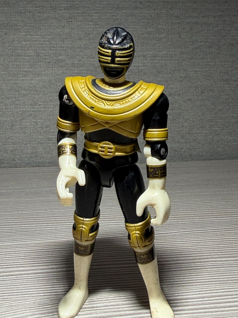 May include: A black and gold Power Ranger action figure. The figure features a black helmet with gold detailing, a gold chest plate, and gold accents on the arms, legs, and belt. The hands and boots are white.