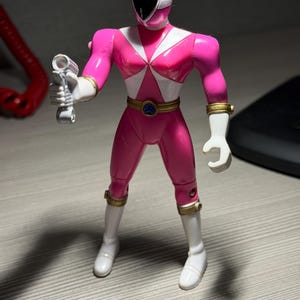 May include: A pink action figure of a Power Ranger, holding a silver weapon. The figure has a white helmet stripe, white accents, and gold wrist and belt details. The figure is wearing white boots and gloves.