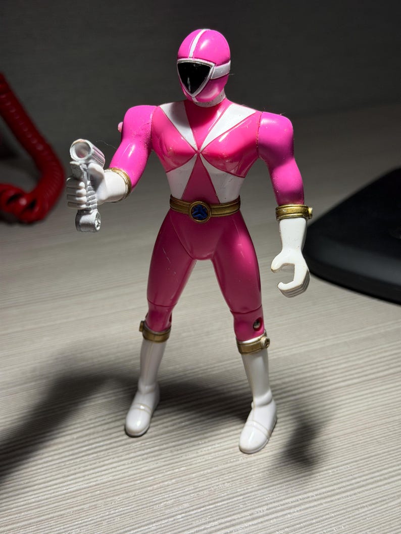 May include: A pink action figure of a Power Ranger, holding a silver weapon. The figure has a white helmet stripe, white accents, and gold wrist and belt details. The figure is wearing white boots and gloves.