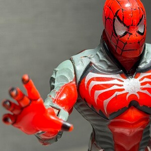 May include: A red and gray Spider-Man action figure with a detailed design. The figure has a red head with a black web pattern and white eyes. The chest features a white spider emblem. The figure is posed with an outstretched hand.