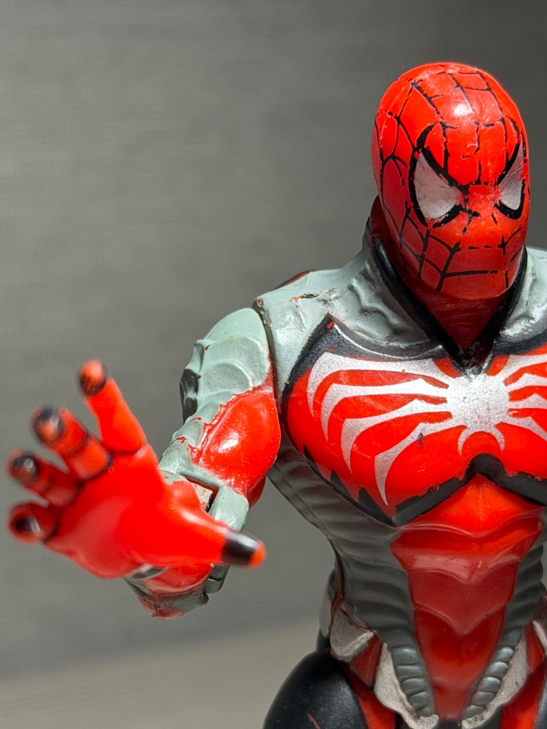 May include: A red and gray Spider-Man action figure with a detailed design. The figure has a red head with a black web pattern and white eyes. The chest features a white spider emblem. The figure is posed with an outstretched hand.