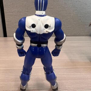 May include: A blue action figure with white accents on the chest and head. The figure has silver details on the arms and legs. The figure is standing with its legs apart.
