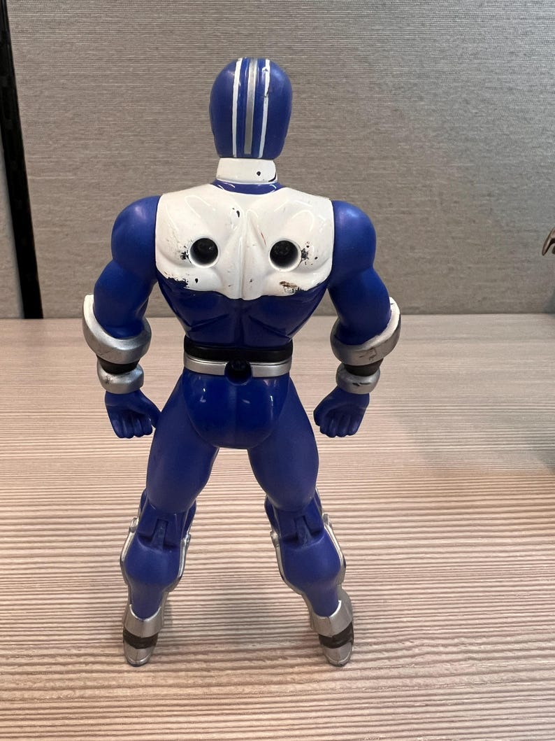 May include: A blue action figure with white accents on the chest and head. The figure has silver details on the arms and legs. The figure is standing with its legs apart.