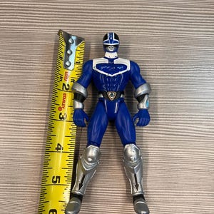 May include: A blue action figure with silver accents, standing upright. The figure is approximately 6 inches tall. It has a helmet, armor on the arms and legs, and a belt with a shield-like emblem. The figure is posed on a light brown surface.