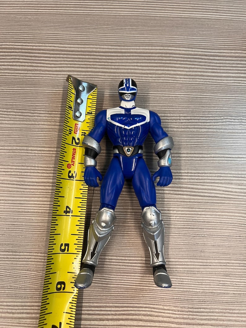 May include: A blue action figure with silver accents, standing upright. The figure is approximately 6 inches tall. It has a helmet, armor on the arms and legs, and a belt with a shield-like emblem. The figure is posed on a light brown surface.