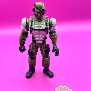 K&ouml;nnte beinhalten: Eine kleine Actionfigur eines Soldaten in Milit&auml;runiform. Die Figur ist haupts&auml;chlich olivgr&uuml;n mit wei&szlig;en und schwarzen Akzenten. Sie hat einen Helm, eine K&ouml;rperpanzerung und Kampfstiefel. Eine M&uuml;nze liegt daneben zum Gr&ouml;&szlig;envergleich.
