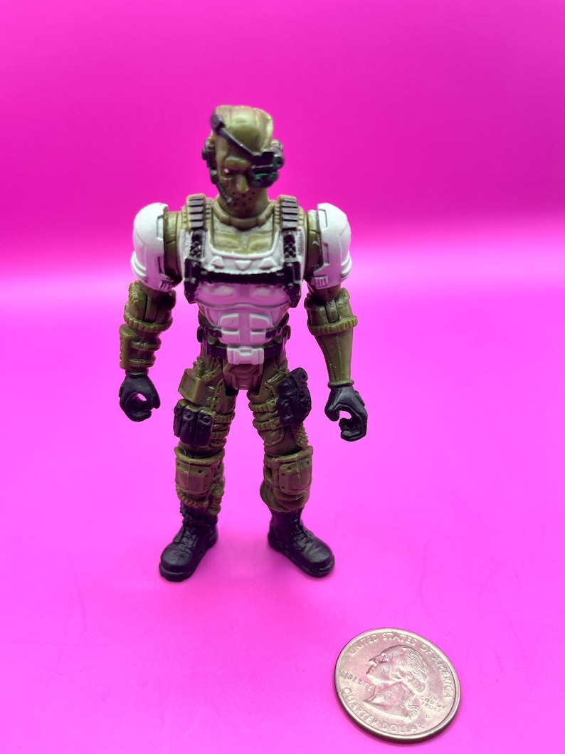 K&ouml;nnte beinhalten: Eine kleine Actionfigur eines Soldaten in Milit&auml;runiform. Die Figur ist haupts&auml;chlich olivgr&uuml;n mit wei&szlig;en und schwarzen Akzenten. Sie hat einen Helm, eine K&ouml;rperpanzerung und Kampfstiefel. Eine M&uuml;nze liegt daneben zum Gr&ouml;&szlig;envergleich.