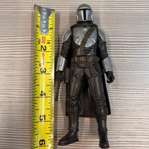 May include: A Mandalorian action figure, approximately 6 inches tall, stands next to a yellow measuring tape. The figure is clad in dark gray and silver armor, holding a blaster pistol. The figure's helmet has a T-shaped visor.