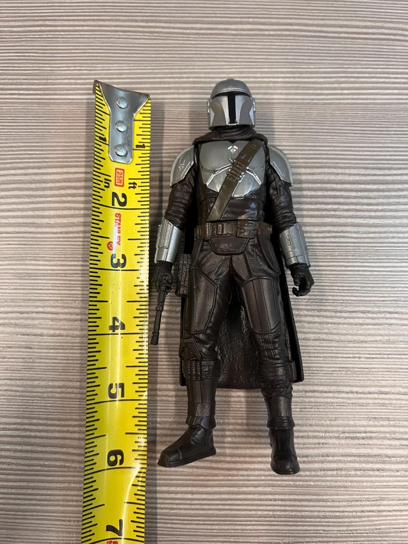 May include: A Mandalorian action figure, approximately 6 inches tall, stands next to a yellow measuring tape. The figure is clad in dark gray and silver armor, holding a blaster pistol. The figure's helmet has a T-shaped visor.