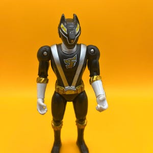 以下が含まれることがあります： 黒とシルバーのパワーレンジャーのアクションフィギュア。オオカミのようなヘルメット、白い手袋、ベルトと手首の袖口に金のアクセントがあります。黄色い背景に立っています。
