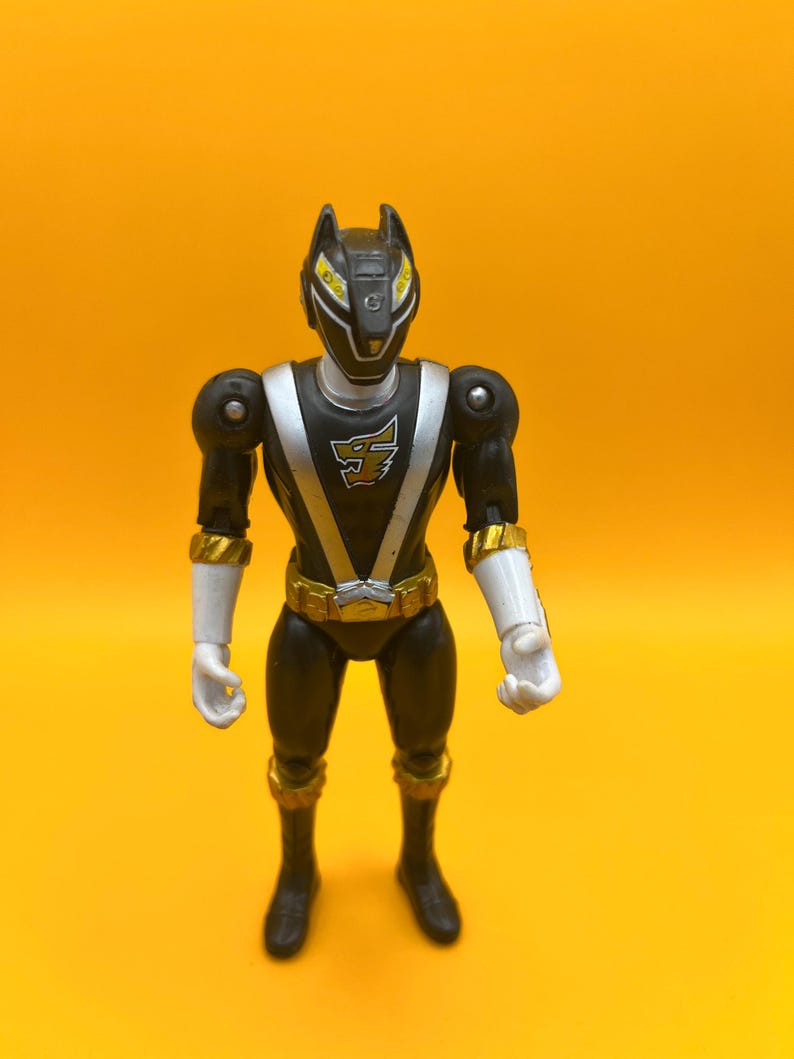 以下が含まれることがあります： 黒とシルバーのパワーレンジャーのアクションフィギュア。オオカミのようなヘルメット、白い手袋、ベルトと手首の袖口に金のアクセントがあります。黄色い背景に立っています。