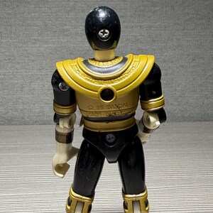 May include: A vintage Power Ranger action figure in black and gold. The figure has a black helmet, gold shoulder armour, and black and gold detailing on the legs and arms. The figure is marked with "&copy; 95 BANDAI MADE IN CHINA."