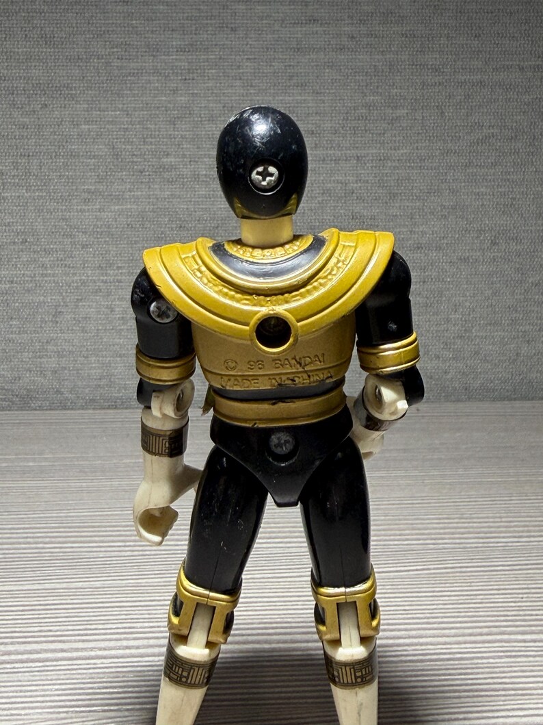 May include: A vintage Power Ranger action figure in black and gold. The figure has a black helmet, gold shoulder armour, and black and gold detailing on the legs and arms. The figure is marked with "&copy; 95 BANDAI MADE IN CHINA."