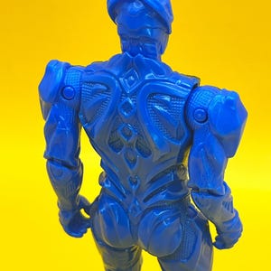May include: A blue action figure, seen from the rear, set against a yellow backdrop. The figure has an intricate, armoured design, including a helmet and sculpted muscles. The figure's pose suggests movement, with arms slightly bent.