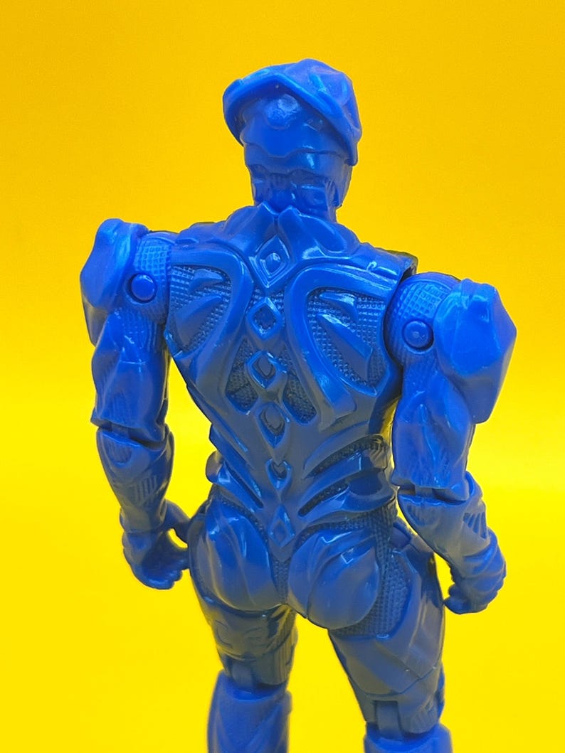 May include: A blue action figure, seen from the rear, set against a yellow backdrop. The figure has an intricate, armoured design, including a helmet and sculpted muscles. The figure's pose suggests movement, with arms slightly bent.