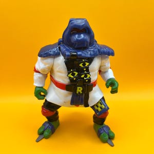 May include: A vintage action figure of a character from the Teenage Mutant Ninja Turtles franchise. The figure is predominantly white with a blue hood and shoulder pads, a black belt with a green and yellow design, and green hands and feet. The figure has a red belt and knee pads.