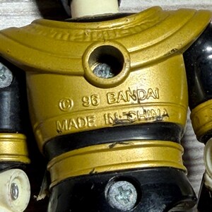 May include: Close-up of a vintage action figure with a gold and black colour scheme. The figure has a gold chest plate with the text "&copy; 96 BANDAI MADE IN CHINA". The figure's joints are black, and the head is black.