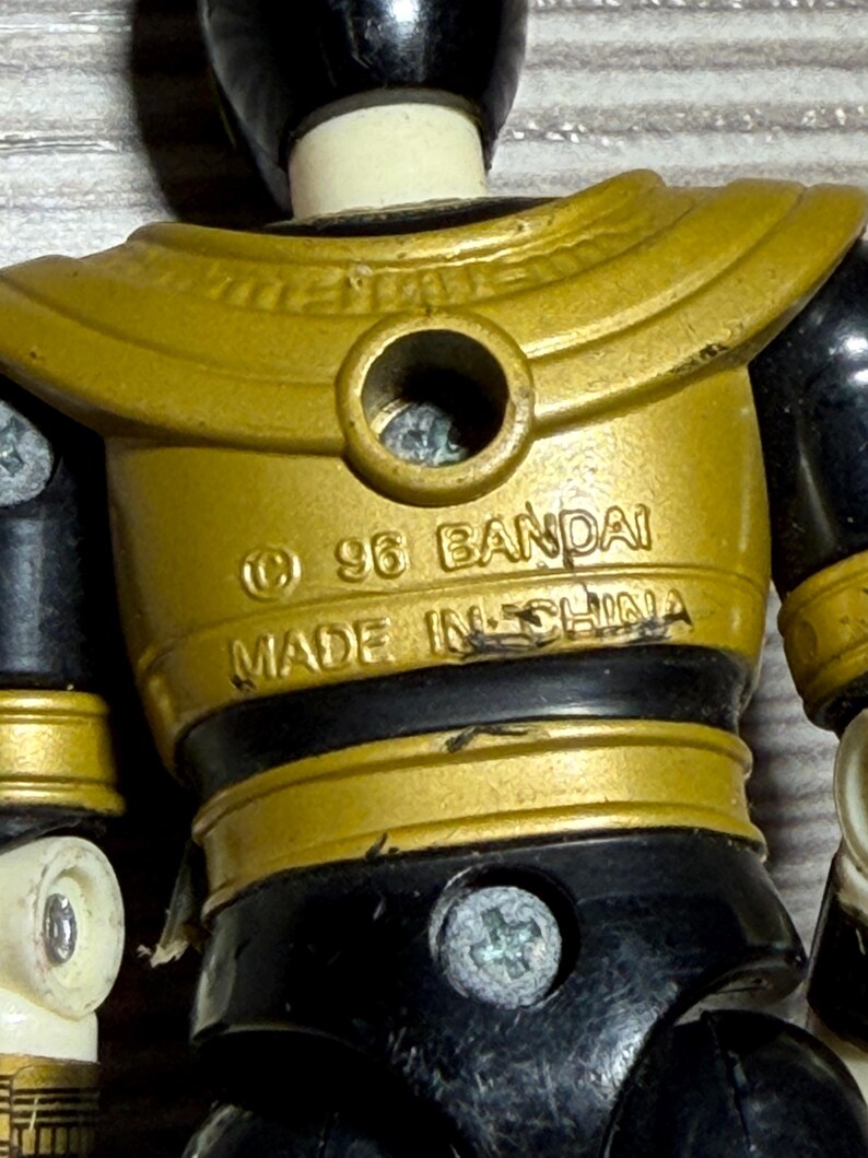 May include: Close-up of a vintage action figure with a gold and black colour scheme. The figure has a gold chest plate with the text "&copy; 96 BANDAI MADE IN CHINA". The figure's joints are black, and the head is black.